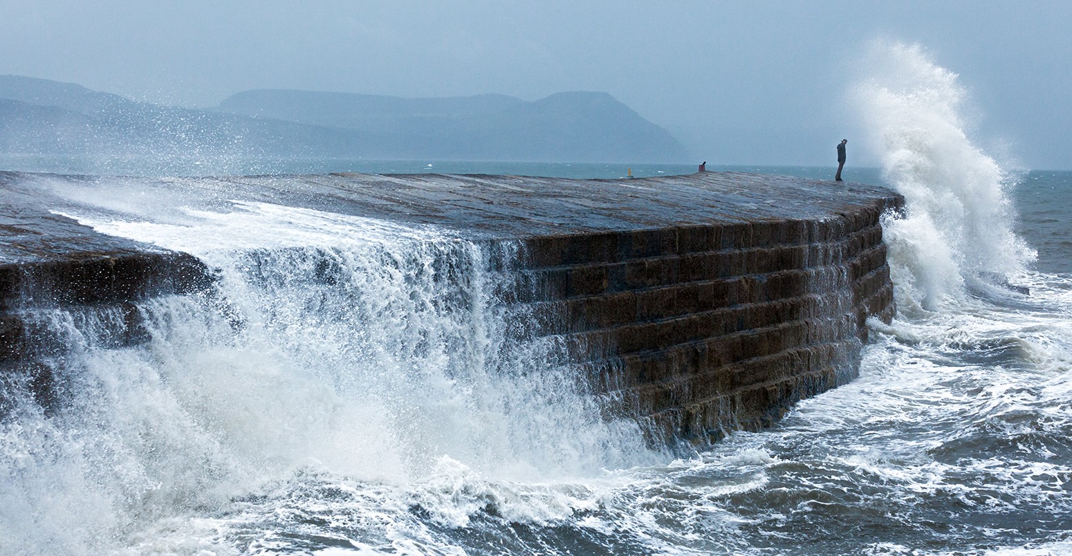 Man standing at the edge of a breakwater and getting a big wave over him.