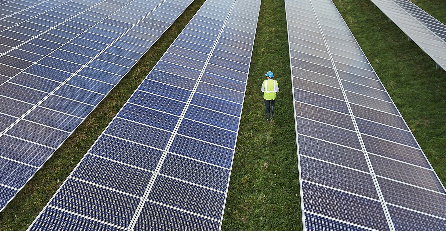 Aerial view of young male Engineer in high vis jacket and blue hard hat surveying a solar farm.