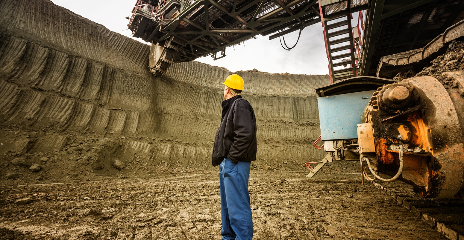 Coal mine worker with a helmet on his head standing in front of huge drill machine and looking at it. Rear view.