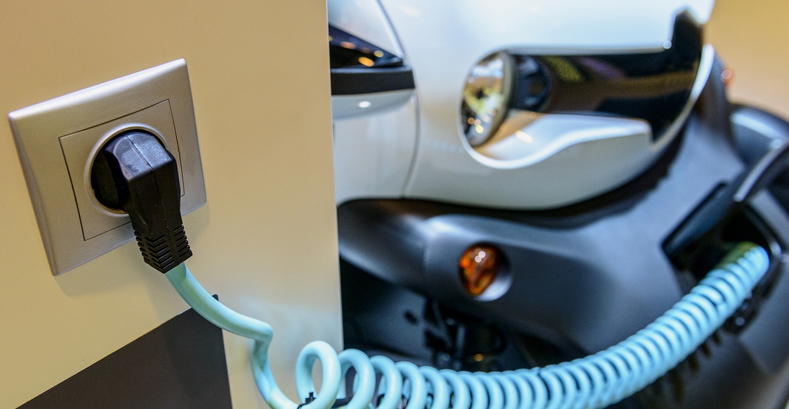 Power supply plugged into an electric vehicle charging station with an electric car in the background.