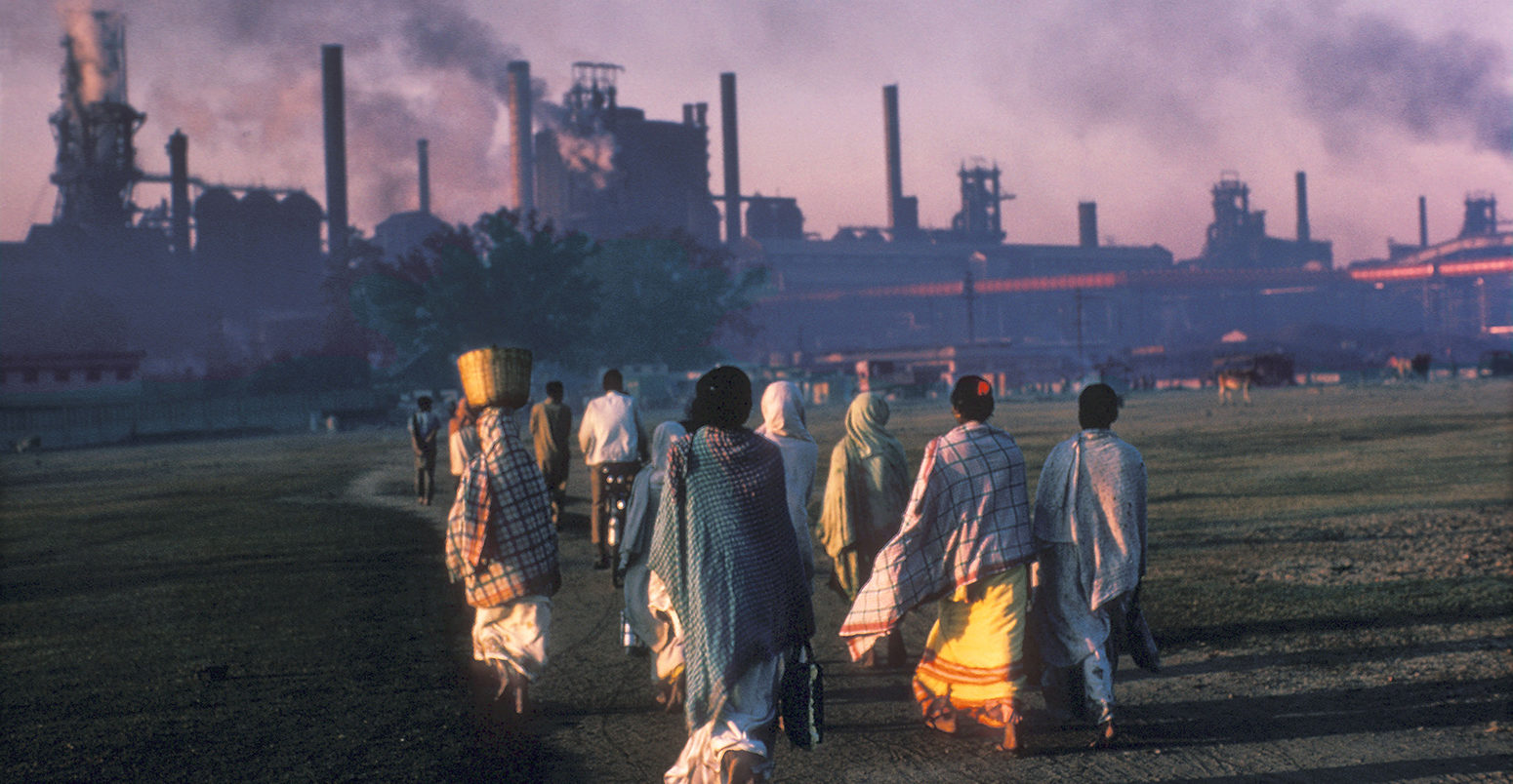 A7PJ6B Indian women in saris with baskets on their heads troop to work at industrial site and power station at sunrise in smoke