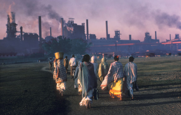 A7PJ6B Indian women in saris with baskets on their heads troop to work at industrial site and power station at sunrise in smoke