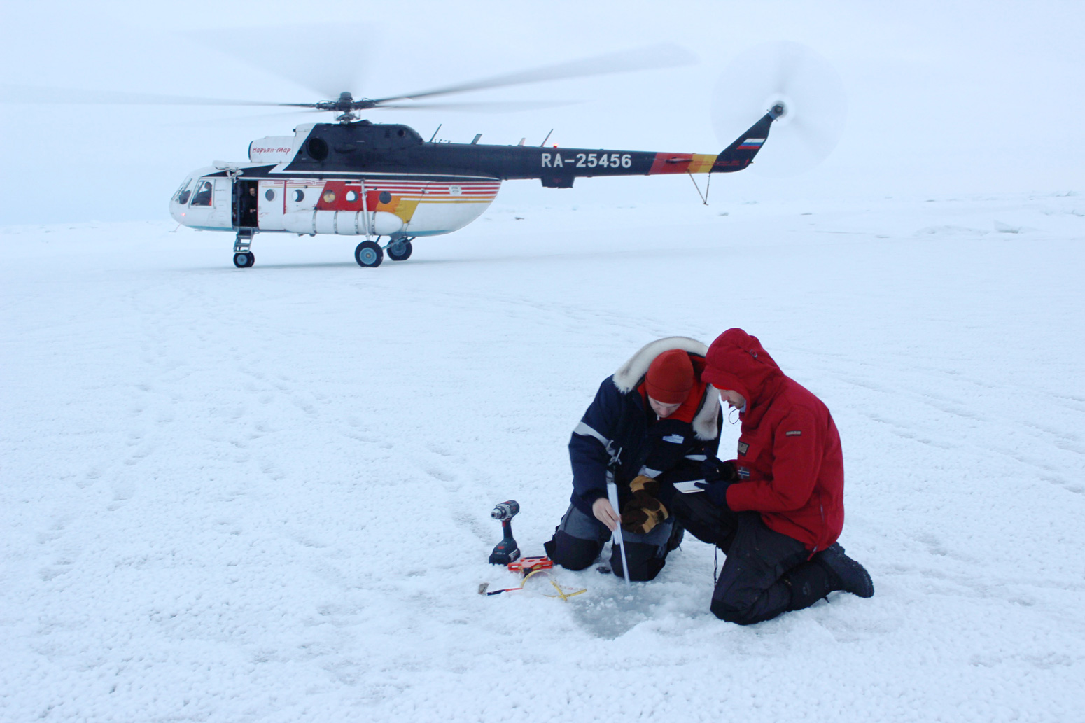 Jan Rohde and Jakob Belter take ice thickness measurements in front of an Mi-8 helicopter in the Central Arctic Ocean. Credit: Daisy Dunne for Carbon Brief. 