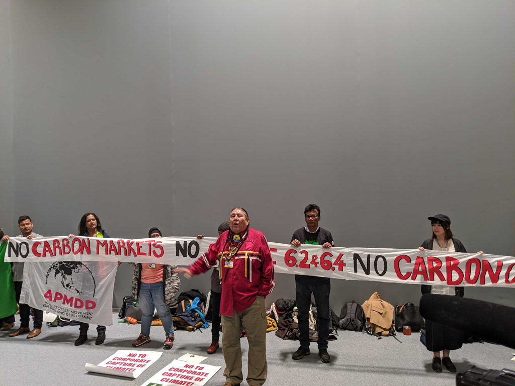 Protestors at COP25 Madrid stand in front of a banner saying "No Carbon Markets, No article 6.2 and 6.4