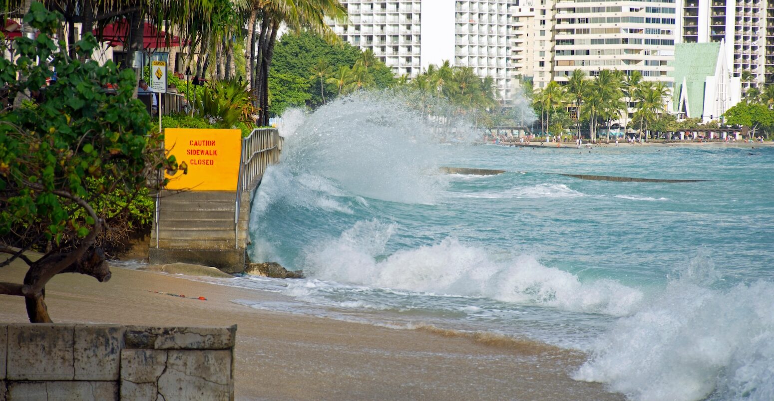 Image shows Waikiki beach in Hawaii, where waves crash against the stairs leading off the beach onto the pavement. A sign at the top of the stairs says 'caution sidewalk closed'. In the background, white high rise buildings.