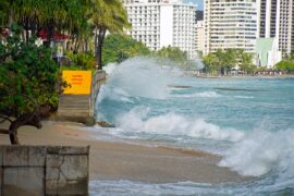Image shows Waikiki beach in Hawaii, where waves crash against the stairs leading off the beach onto the pavement. A sign at the top of the stairs says 'caution sidewalk closed'. In the background, white high rise buildings.