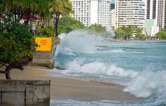 Image shows Waikiki beach in Hawaii, where waves crash against the stairs leading off the beach onto the pavement. A sign at the top of the stairs says 'caution sidewalk closed'. In the background, white high rise buildings.