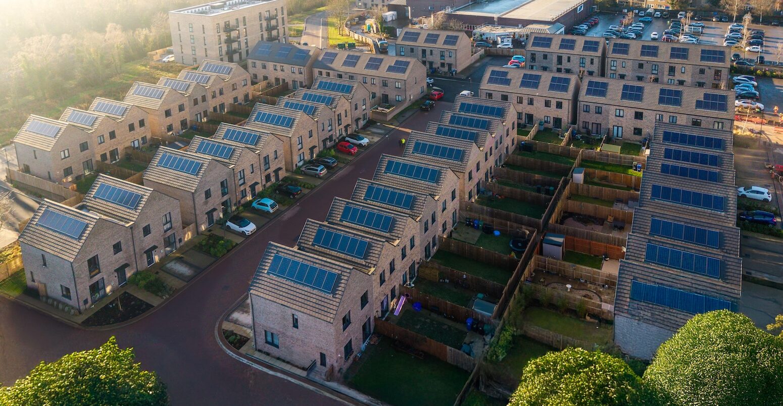 An almost birds-eye view of houses. Rows of new-build houses going from the bottom left hand corner to the middle of the image. More houses are in rows perpendicular to this, in the top right hand corner of the photo. All houses have roof-topped solar, a garden and a drive.