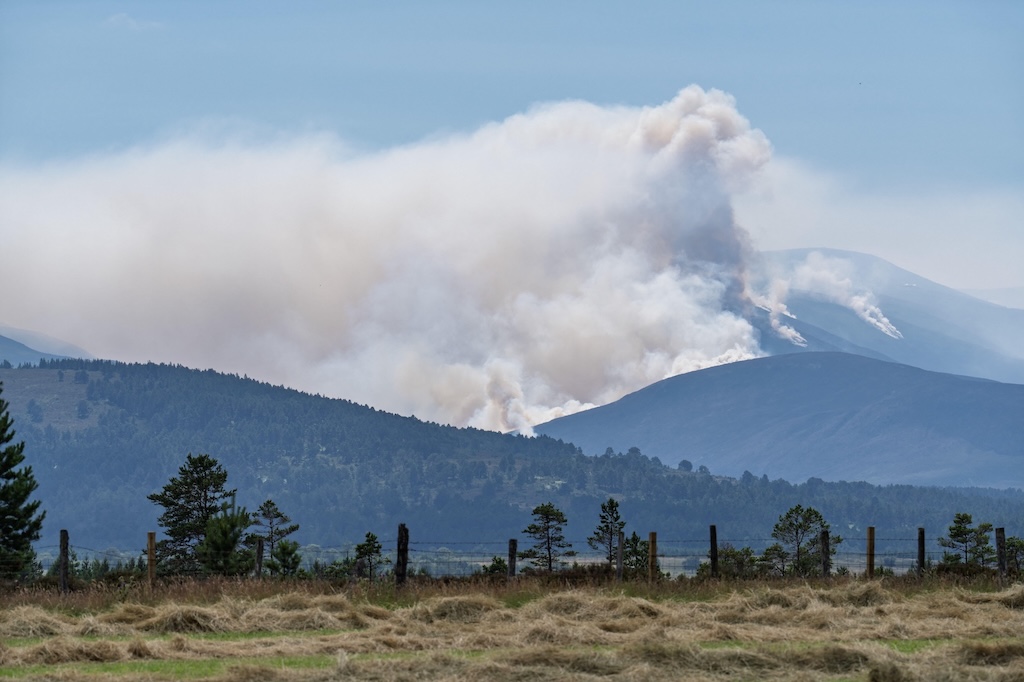 Active wildfire (wildfire itself not visible in the photo) producing thick white smoke over blue layers of forested mountains, with a dry yellow and green meadow, and rustic fence dotted with a handful of green trees, in the foreground. All under a clear blue sky.